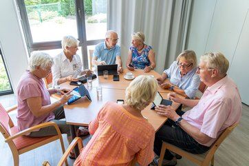 Bild mit fünf Seniorinnen und zwei Senioren, die mit Tablets rund um einen Tisch sitzen, sich unterhalten und damit arbeiten