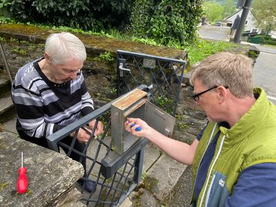 Mann schraubt Briefkasten am Eingangstor fest, alte Frau steht hinter dem Tor und schaut zu.