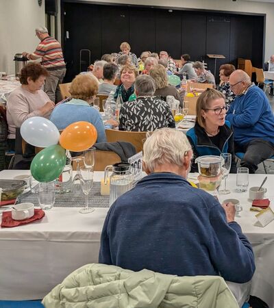 Viele gedeckte und geschmückte Tische, an denen Männer und Frauen unterschiedlichen Alters in einem größeren Raum sitzen, sich unterhalten, essen und trinken