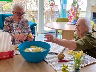 2 alte Damen sind über Eck am Tisch und schälen Kartoffeln, die geschälten Kartoffeln schwimmen in einer blauen Schüssel mit Wasser auf der Tischmitte.
