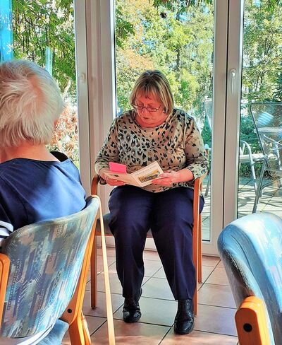 Frau über Buch gebeugt beim Vorlesen vor einer Gruppe älterer Menschen