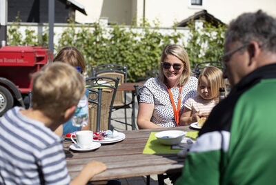 Ein Mann und drei Kinder sitzen mit Frau Zinken an einem Holztisch mit Kaffegeschirr im Freien