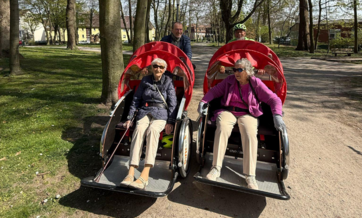 Bild mit zwei ältern Damen mit Sonnenbrillen, die in Rikschas von zwei Männern durch einen Park gefahren werden.