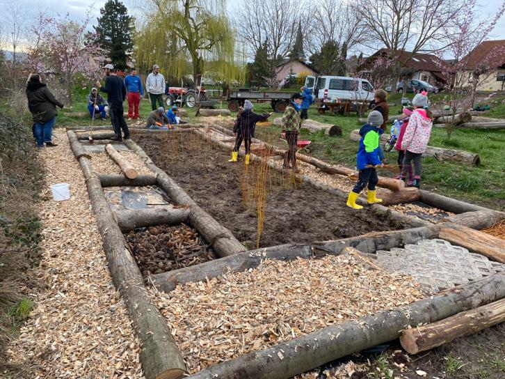 Kinder, Männer und Frauen warm angezogen im Frühling. Auf dem Boden liegen Baumstämme die einen Parcours abstecken, verschiedene Räume sind mit Holzspänen, Steinen, Erde und anderen Materialien gefüllt