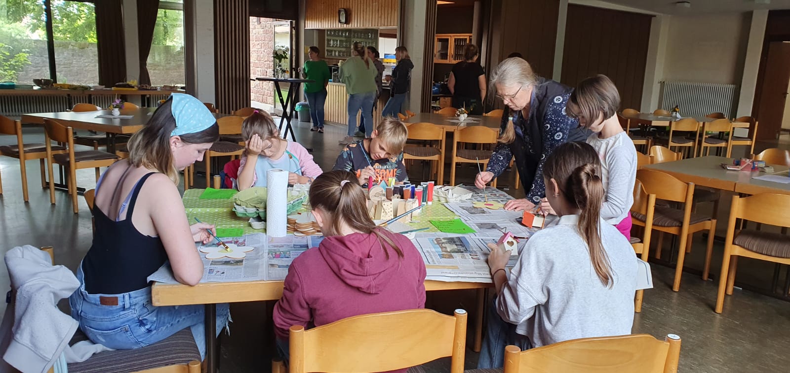 fünf Kinder und zwei Frauen sind an einem Gruppentisch mit Malarbeiten beschäftigt.