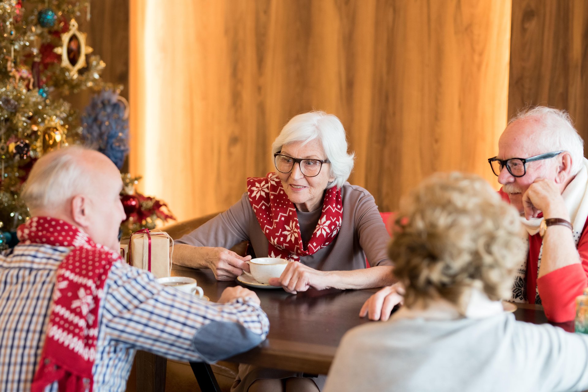 Bild mit zwei älteren Frauen und Männern, die Schals mit Weihnachtsmotiven tragen. Sie sitzen sich unterhaltend gemeinsam an einem Tisch mit Cafeetassen und Geschenken, im Hintergrund ein Weihnachtsbaum