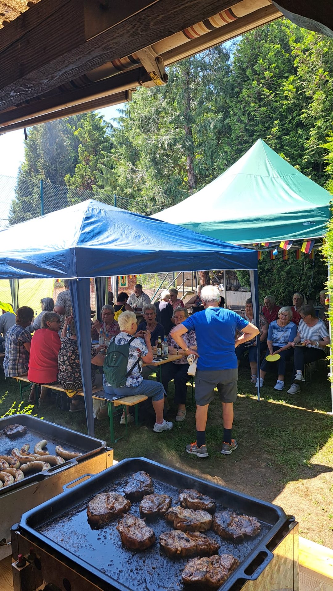 auf zwei Grills liegen Steaks und Würstchen, dahinter sind Zelte aufgebaut in denen Ältere in Sommerkleidung sitzen und stehen