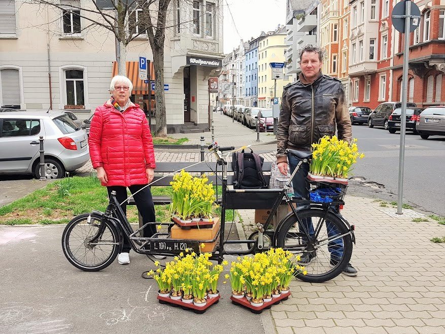 Frau und Mann mit stehen am Lastenrad, das mit Paletten gelb blühender Osterglocken beladen ist.