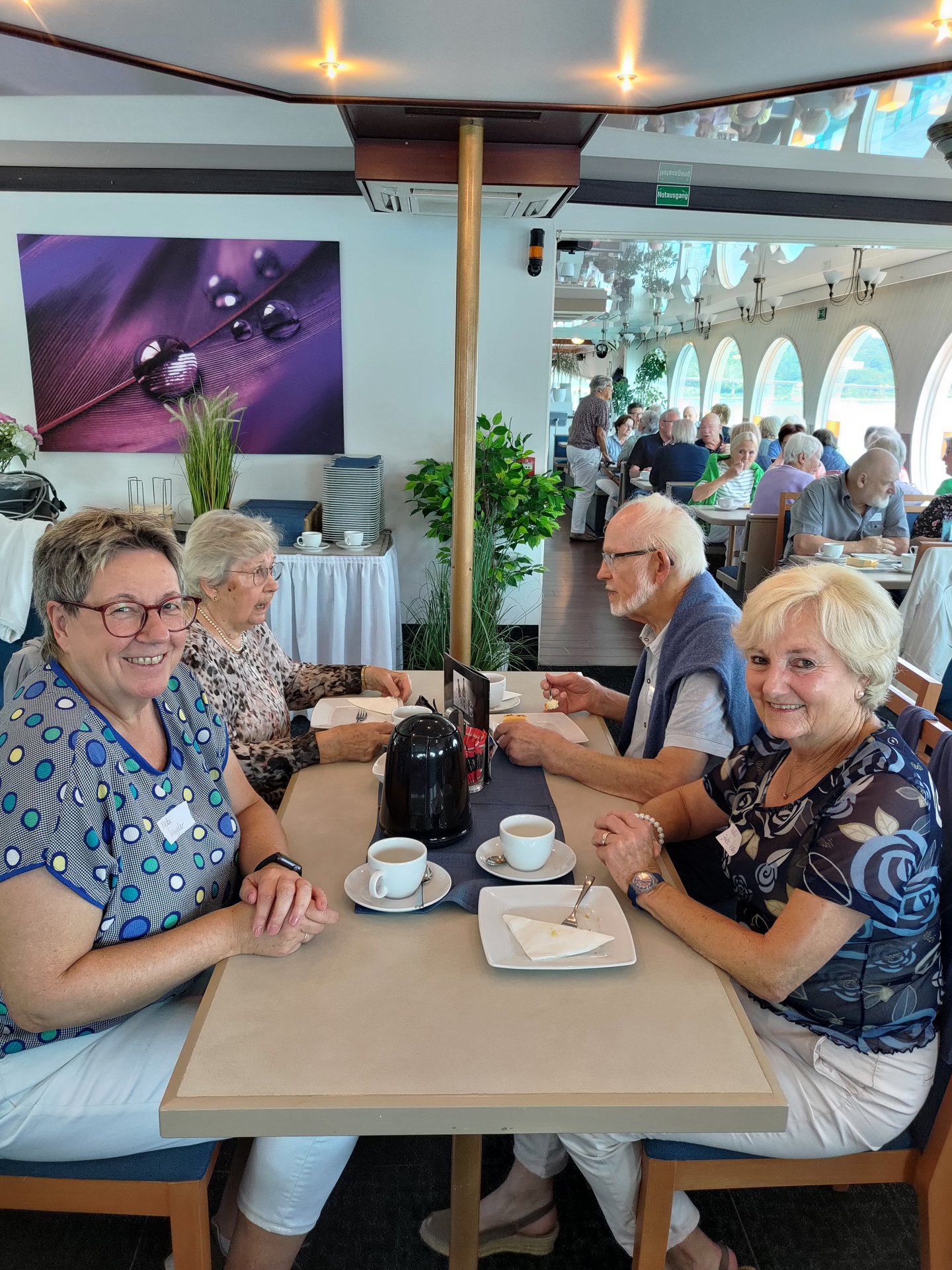 Bild mit älteren Frauen und Männern, die mit Kaffee an mehreren Tischen auf einem Ausflugsschiff sitzen.
