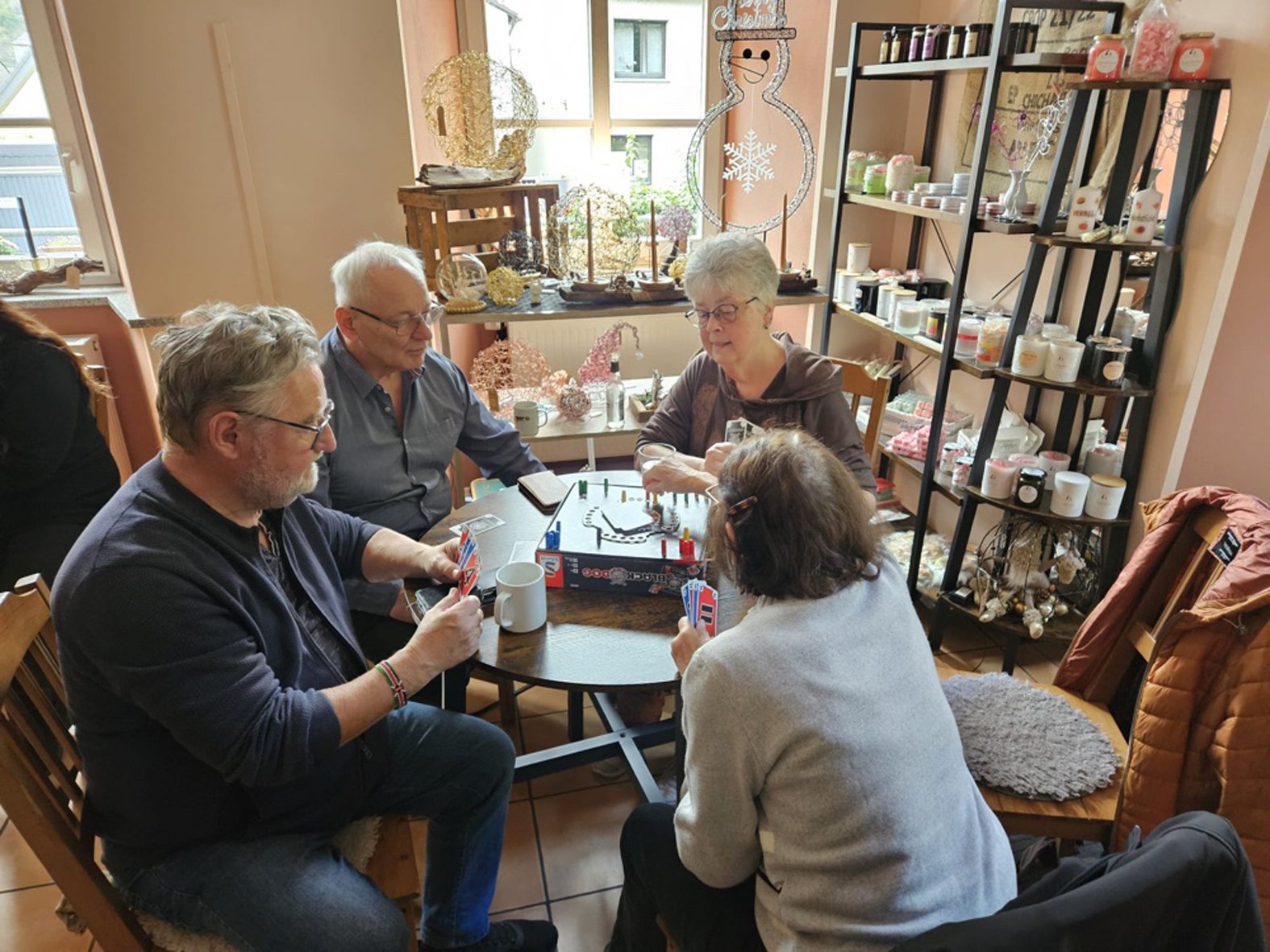 Bild mit je zwei älteren Männern und Frauen an einem Tisch im Ladeninterieur mit Regealen. Sie halten Karten in der Hand auf der Tischmitte ein Spielbrett sowie Tassen.