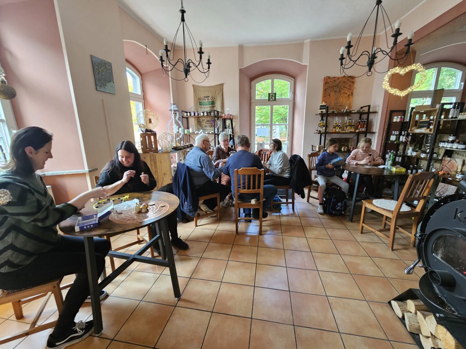Bild mit Blick in einen Ladenraum, in dem sich mehrere Tische befinden an denen Personen unterschiedlichebn Alters verschiedene Spiele spielen, im Vordergrund ein Holzofen