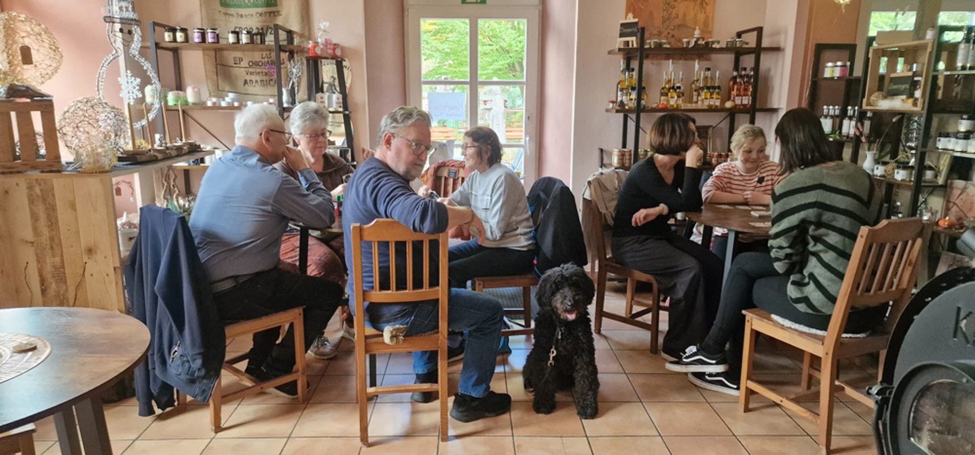 Bild aus einem Ladengeschäft in dem zwei Tische stehen um die herum Männer und Frauen beim Spiel sitzen, ein Hund im Vordergrund
