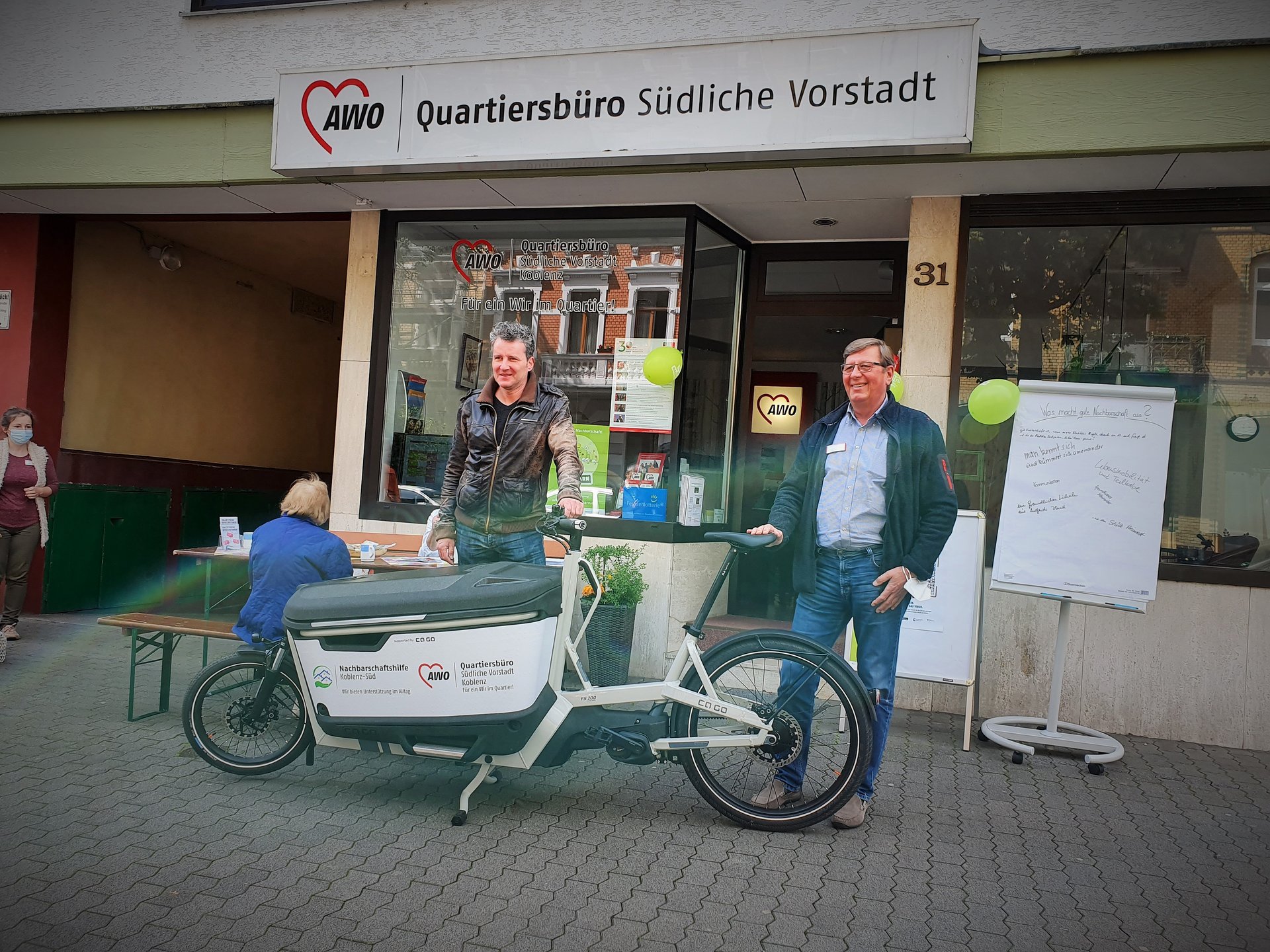 2 Männer stehen hinter elektrischem Lastenrad auf der Straße vor dem Quartiersbüro