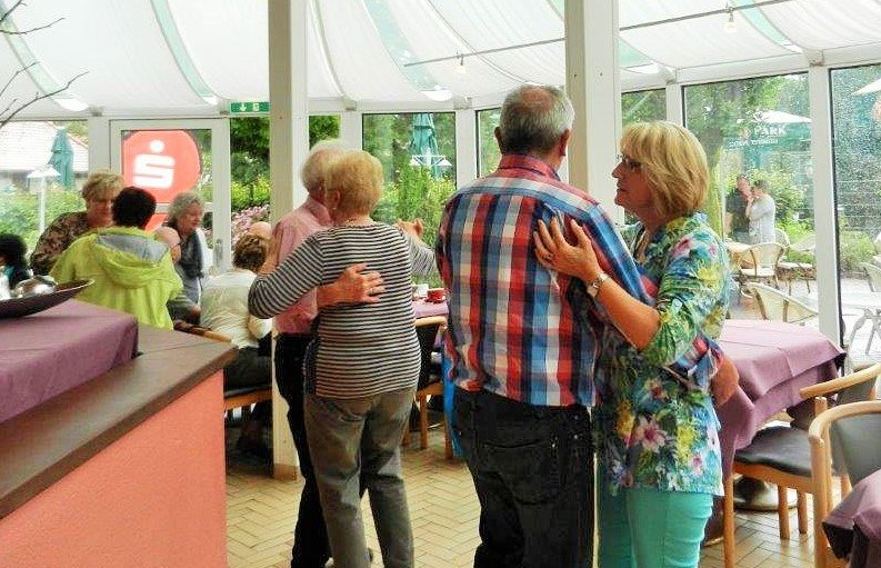 zwei tanzende Paare in einem Wintergarten, im Hintergrund sitzen Menschen an einer Bar und an Tischen und unterhalten sich
