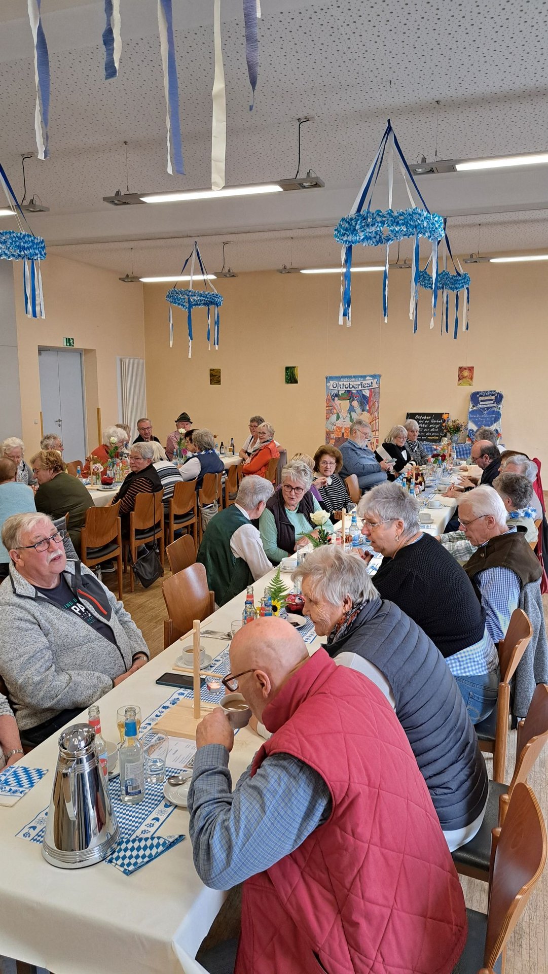 an langen Tischtafeln die bayerisch blau-weiß geschmückt sind sitzen Senioren beim Speisen und Getränken. In der Halle hängen einige blau-weiße Kränze von der Decke