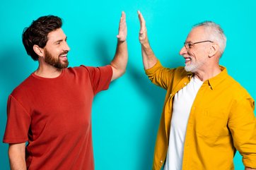 Bild mit einem jungen und alten Mann, die lachend und mit erhobenen Armen ihre Handflächen zusammenschlagen.