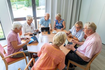 Bild mit fünf Seniorinnen und zwei Senioren, die mit Tablets rund um einen Tisch sitzen, sich unterhalten und damit arbeiten