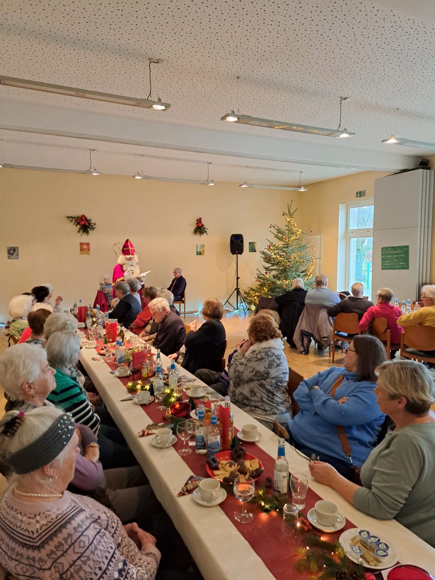 Senior*innen sitzen an langen weihnachtlich geschmückten Tafeln mit Kerzen und hören einem Nikolaus zu, daneben steht ein Weihnachtsbaum